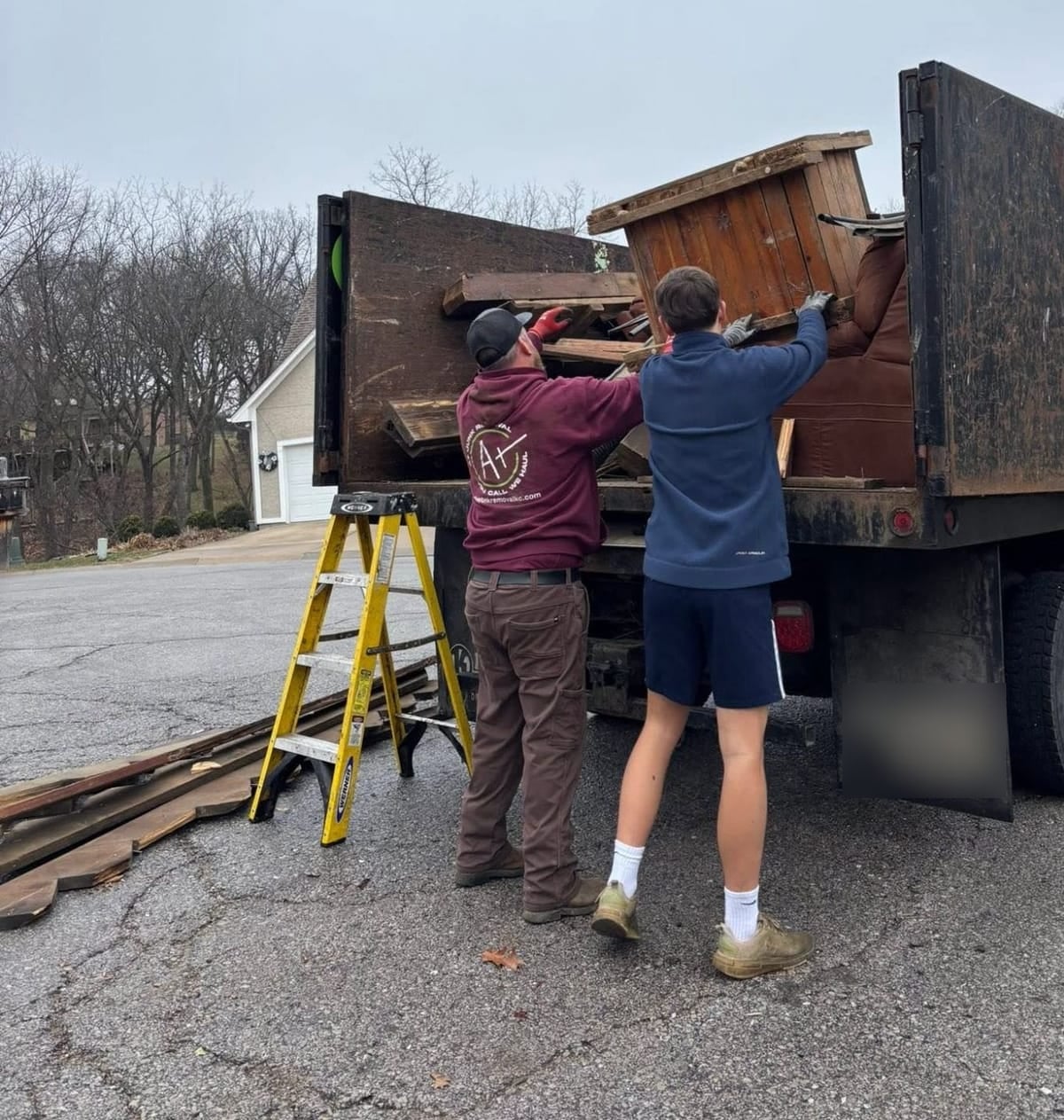Two A Plus crew members lifting old wood furniture into the dump truck in a driveway